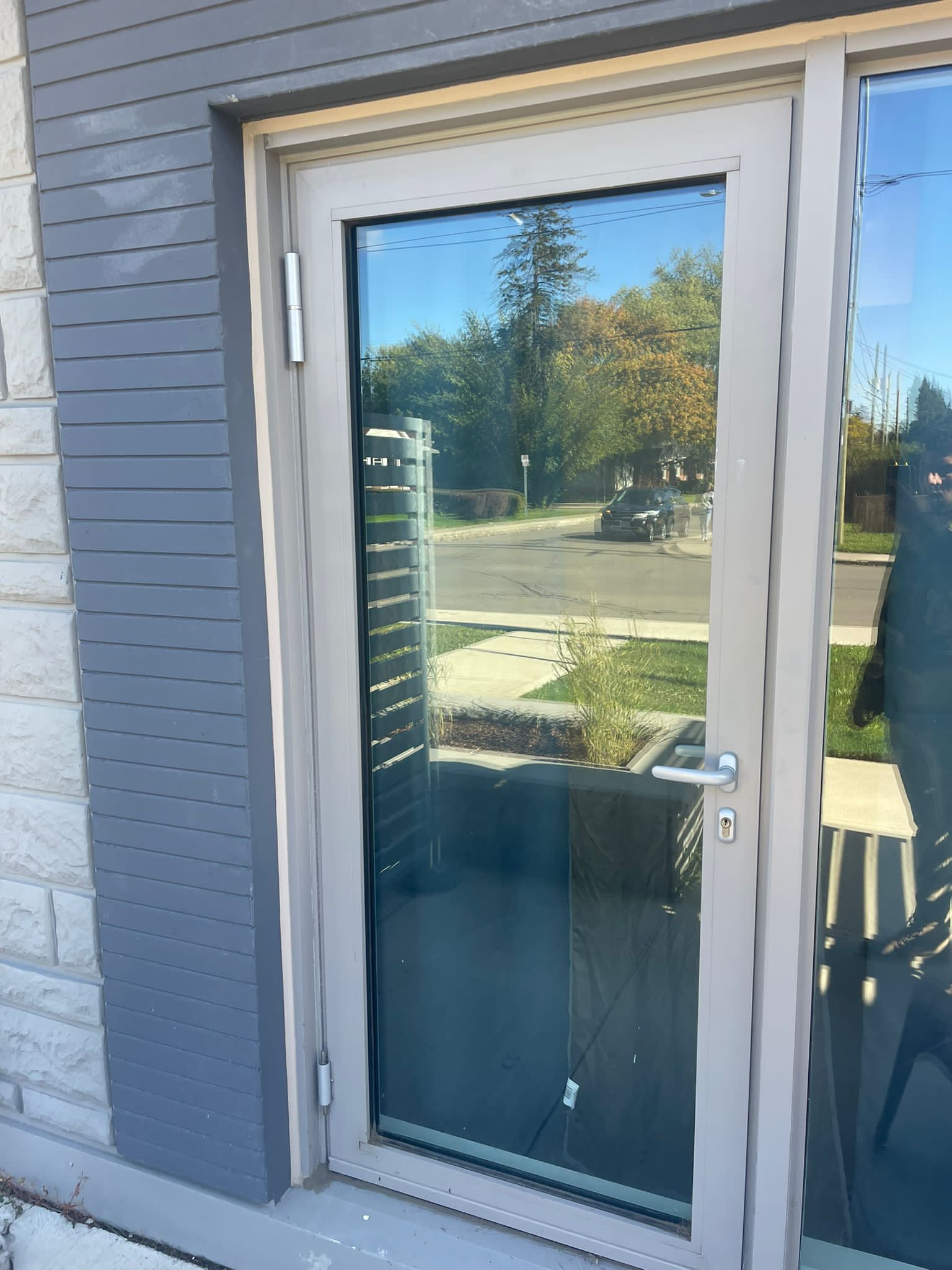 Modern glass door with light gray frame reflecting a street scene, trees, and blue sky.